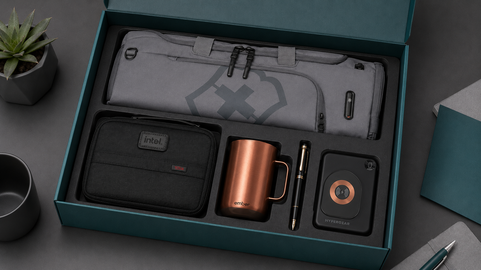 Executive employee welcome kit with a gray duffel bag, Intel tech pouch, copper Ember mug, black pen, and foldable wireless charging stand arranged in an EPI-themed teal gift box.