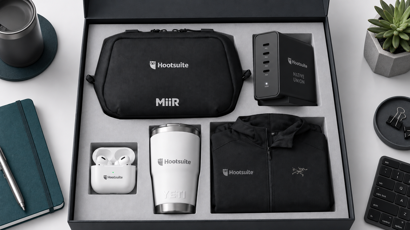 Hootsuite branded IT team welcome kit with a black tech pouch, multi-port charger, wireless earbuds, YETI tumbler, and black jacket arranged in a premium gift box.