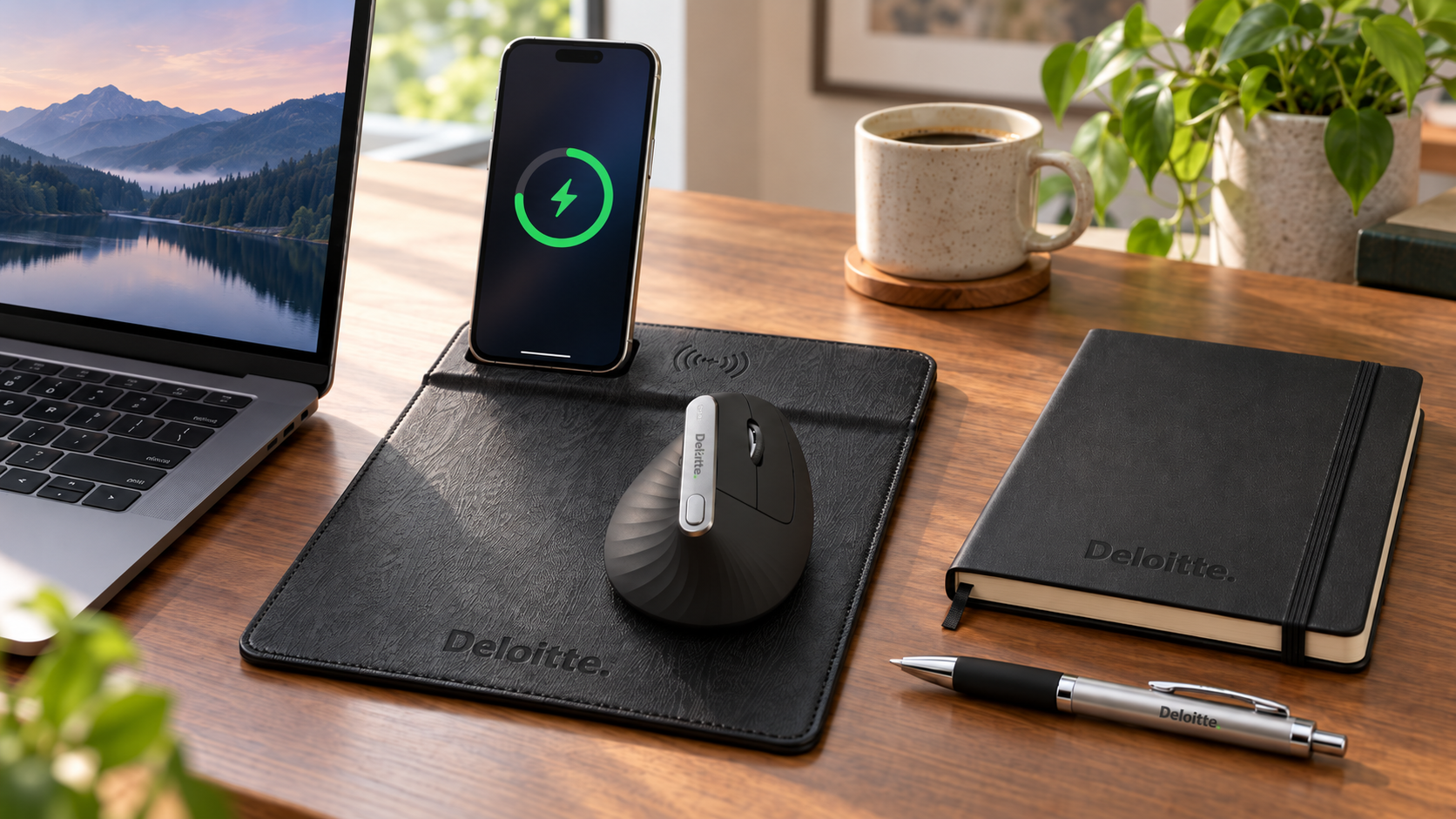 Best remote employee gifts for a home office featuring a black Deloitte branded wireless charging mouse pad with a smartphone charging upright, a black Logitech MX Vertical ergonomic mouse, a black Moleskine notebook, and a Deloitte branded pen beside a laptop, coffee mug, and green plants on a wooden desk.