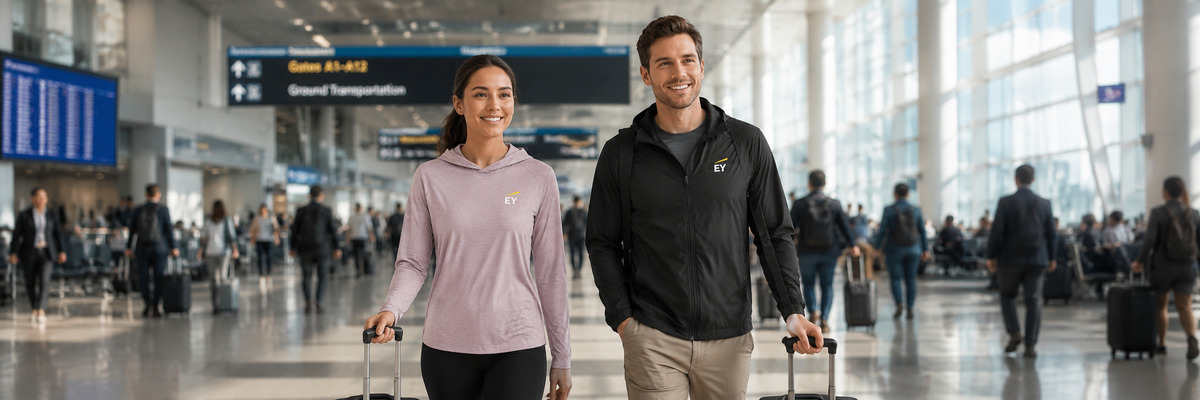 Patagonia corporate apparel worn by two travelers walking through a busy airport terminal with rolling luggage, featuring an EY-logo-branded Patagonia Men's Houdini Jacket and an EY-logo-branded Patagonia Ladies' Capilene Cool Daily Hoody.