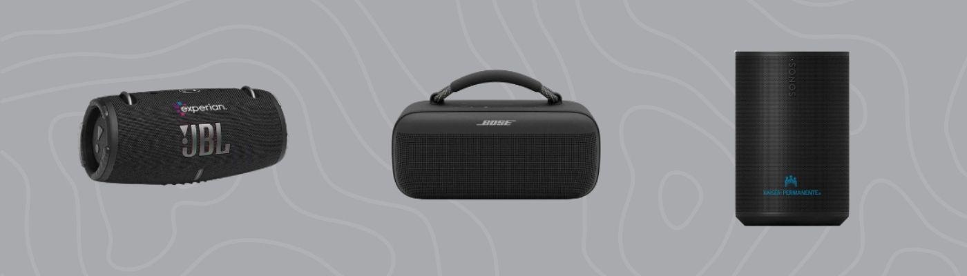 A lineup of portable tech gifts, including JBL, Bose, and Sonos wireless speakers, is displayed on a grey, patterned background.